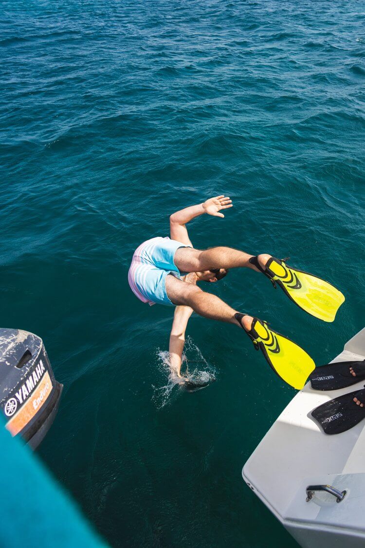 Adult dives headfirst into turquoise water from a boat wearing fins; splashes visible. Yamaha engine and hull visible.