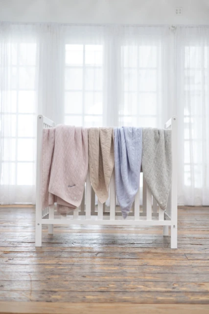 Four pastel-colored, knitted baby blankets hanging on a white crib against a backdrop of bright windows and light wood flooring.