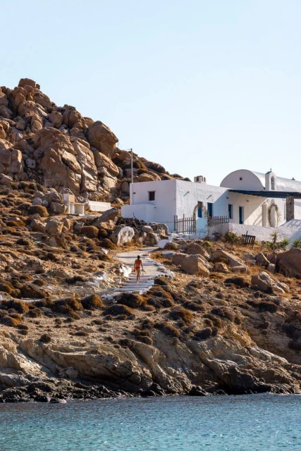 Person walks up white stone steps toward a whitewashed building, set against a rocky hillside and blue sky with visible ocean.