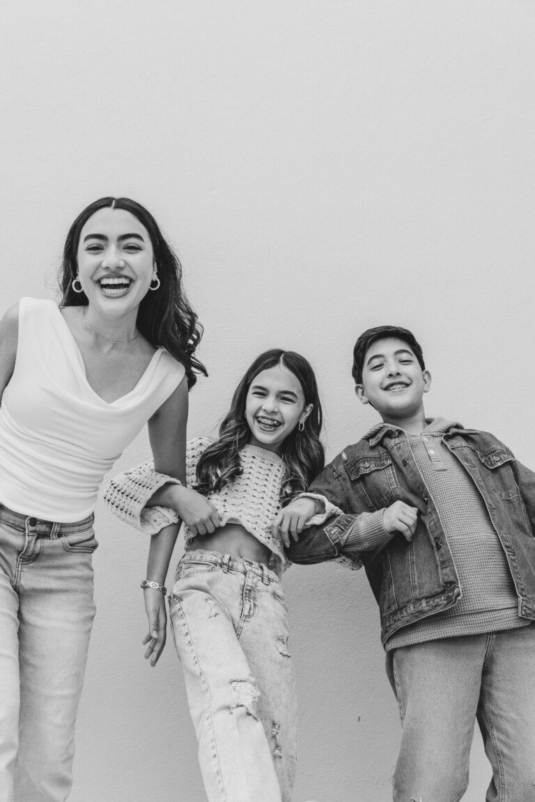 Black and white shot of a smiling family posing in front of a wall: an adult woman, a teen girl, and a teen boy standing close together.