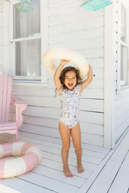 A smiling child in a swimsuit holds a floatation ring above their head on a porch, with a beachy, summery vibe and natural lighting.