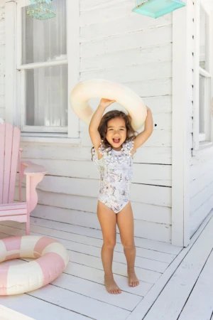 A smiling child in a swimsuit holds a floatation ring above their head on a porch, with a beachy, summery vibe and natural lighting.