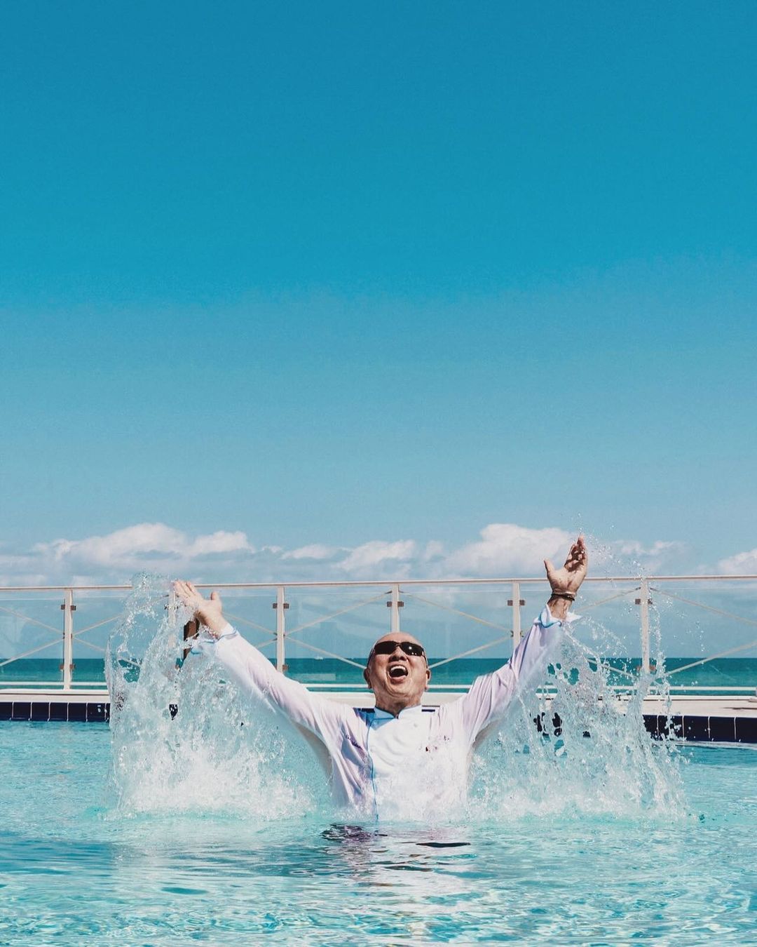 Middle-aged man with sunglasses jumps in a pool, arms raised in excitement against a backdrop of a turquoise sea and sunny sky.