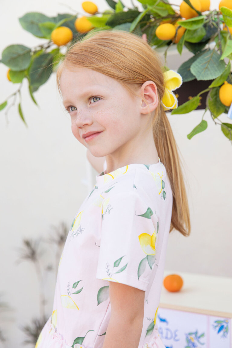 Portrait of a young girl with red hair wearing a dress with a lemon pattern, standing in front of a refreshment stand.