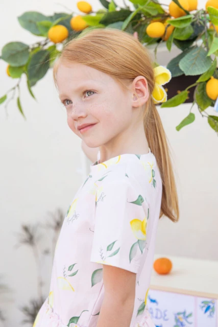 Portrait of a young girl with red hair wearing a dress with a lemon pattern, standing in front of a refreshment stand.