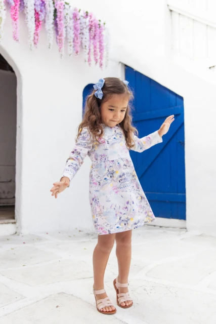 A little girl in a white dress with a blue bow stands in a white-walled courtyard with a blue door and hanging purple flowers.