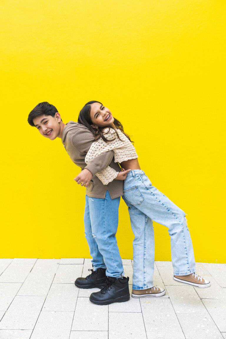 Two smiling teenagers lean back-to-back against a vibrant yellow wall outdoors, wearing casual clothing and dark shoes on a tiled surface.