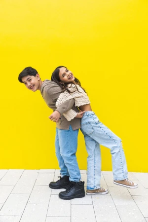 Two smiling teenagers lean back-to-back against a vibrant yellow wall outdoors, wearing casual clothing and dark shoes on a tiled surface.