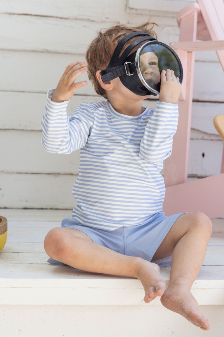 A young child sits on a white porch wearing a striped shirt and shorts with a large diving mask covering their face.
