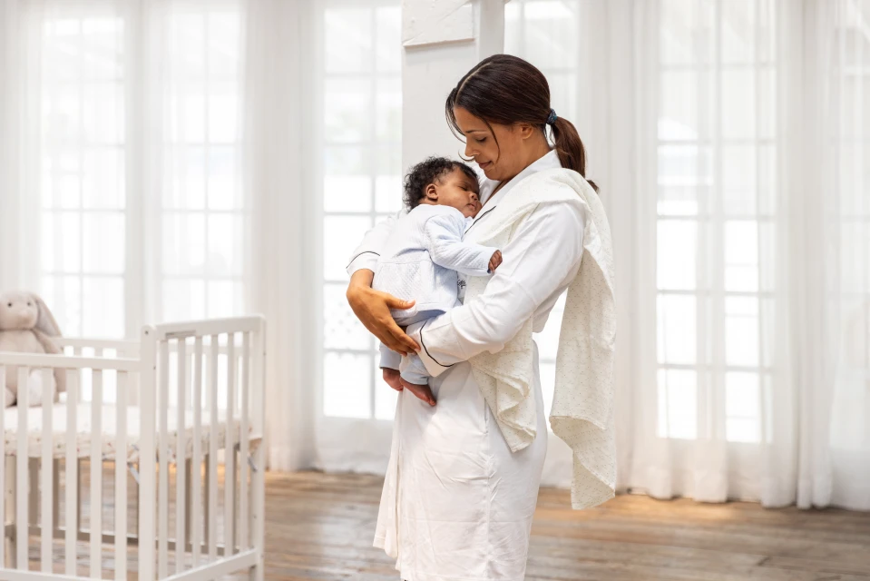 A woman in a white robe is indoors holding a sleeping baby in her arms, next to a baby crib, with large white windows in the background.