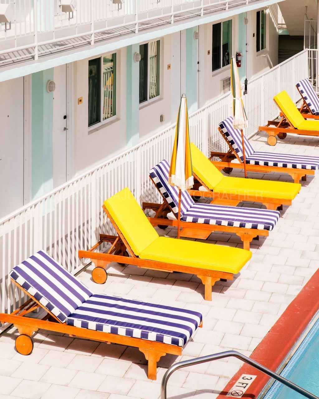 Row of beach chairs with striped cushions and yellow umbrellas next to a swimming pool and a building with white walls.