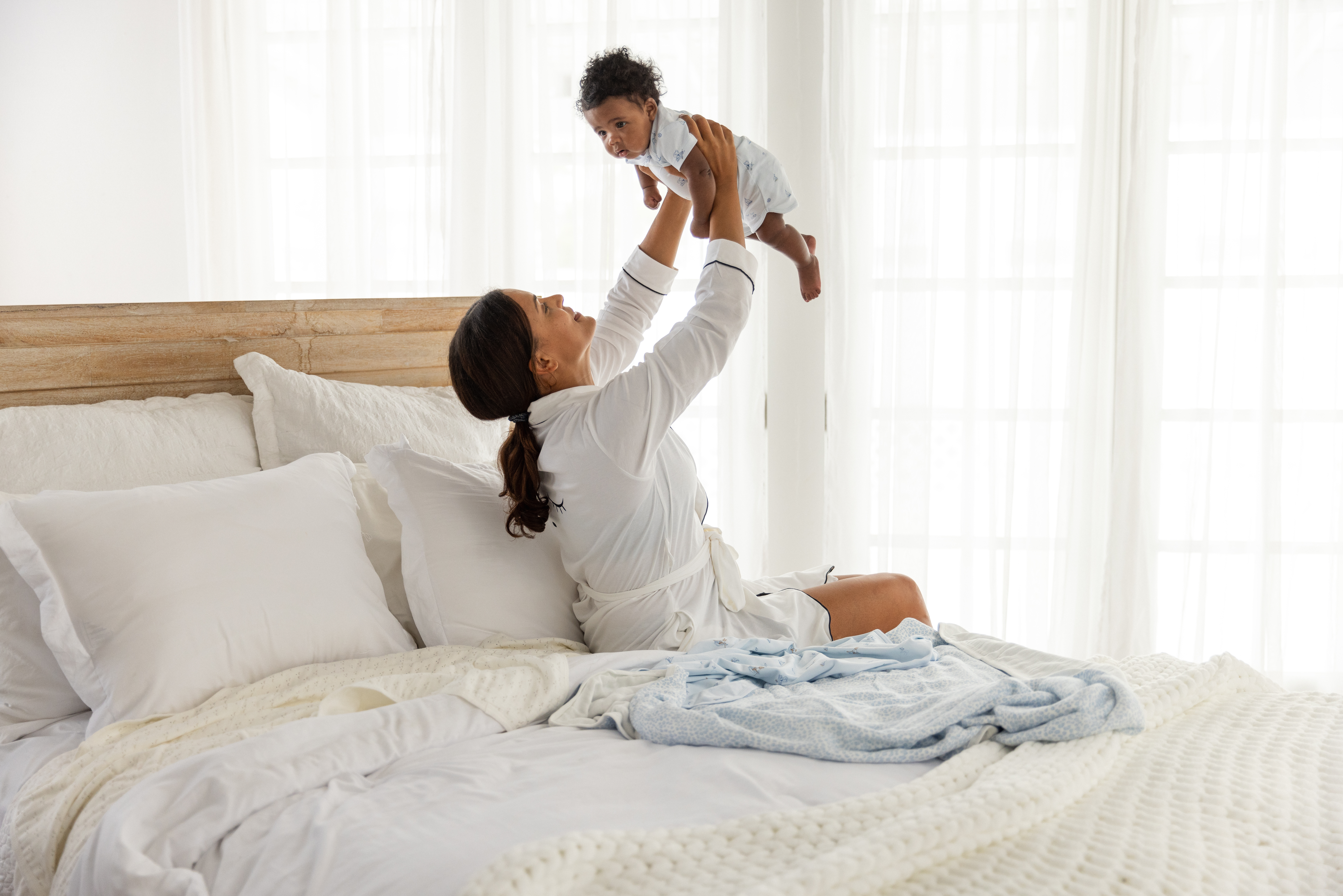 Smiling woman in white robe holding baby in the air above a white bed in a brightly lit bedroom near white curtains.