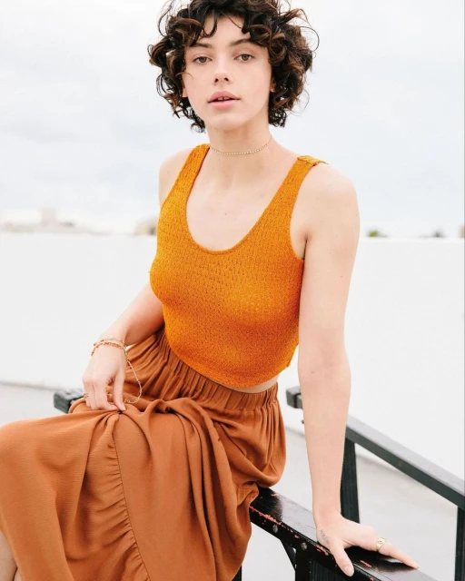 Portrait of a young adult female sitting on a black metal railing, wearing a brown skirt and orange top, with a blurred white background.