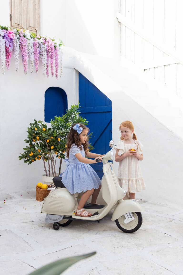 Two young girls posing with a toy Vespa scooter in front of a whitewashed wall with blue door and flower garlands and lemon tree.