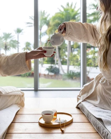 Tea service at a tranquil spa: two women in robes, one pouring tea into a cup by a large window overlooking lush tropical greenery.