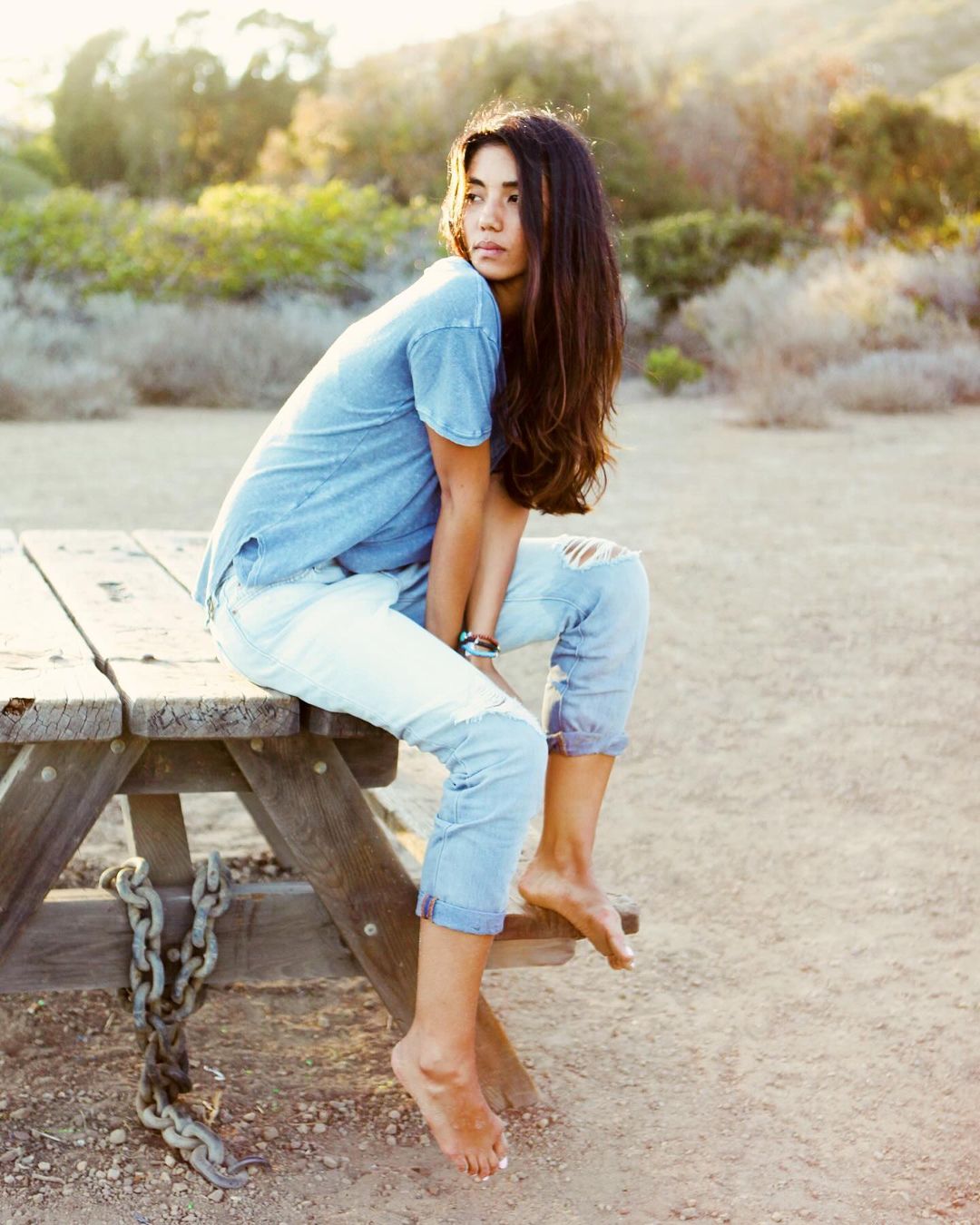 Young woman in denim shirt and ripped jeans sits barefoot on a wooden picnic table in a sunlit, sandy outdoor environment.