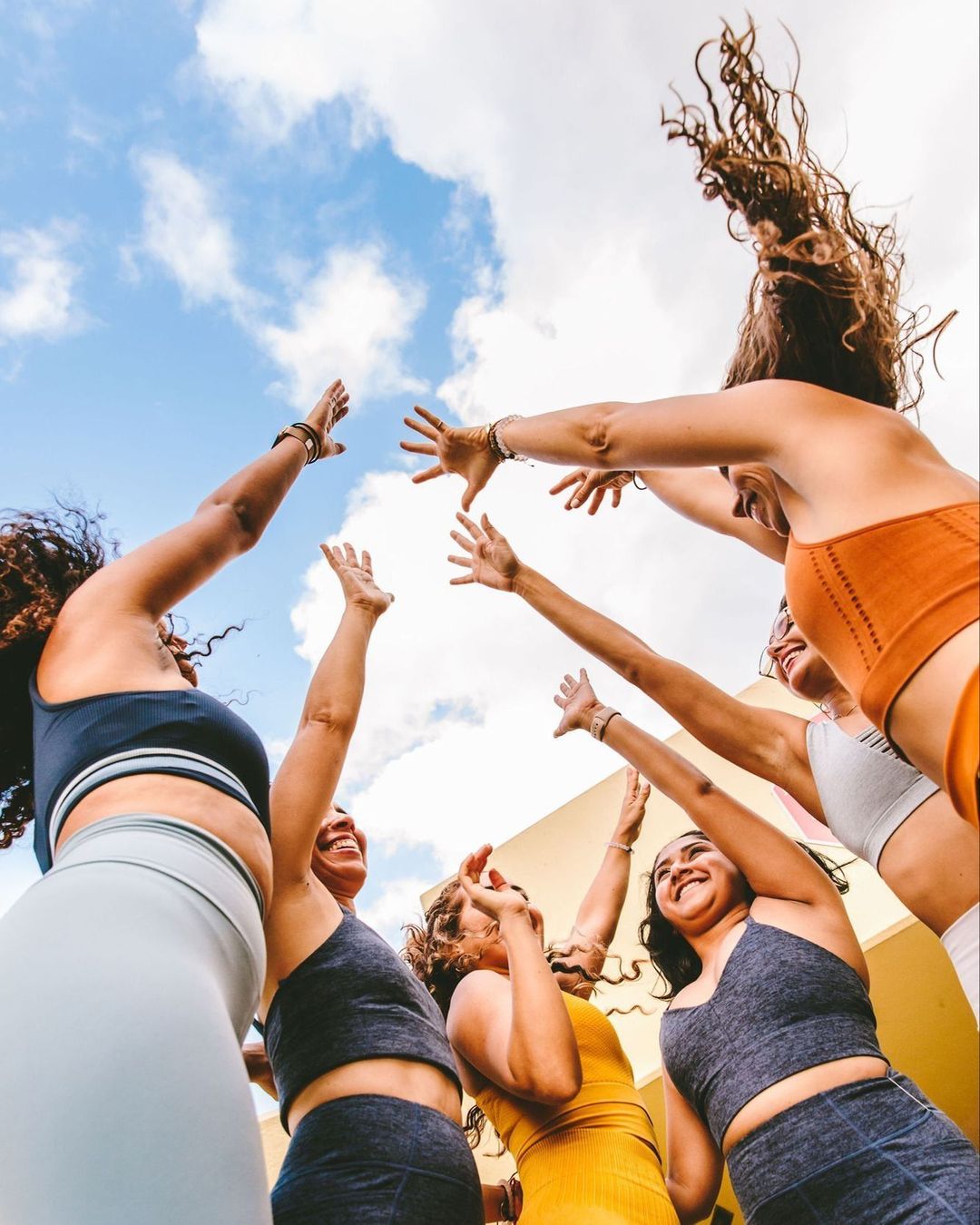 A group of six adult women in athletic wear stand outside reaching their hands towards a blue, partly cloudy sky in a low-angle shot.