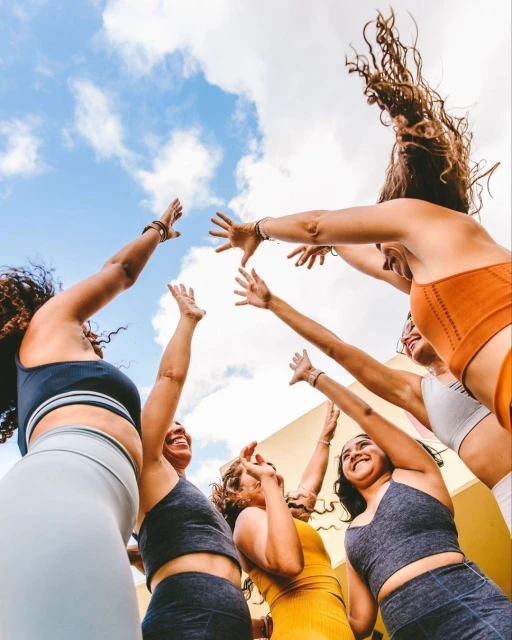 A group of six adult women in athletic wear stand outside reaching their hands towards a blue, partly cloudy sky in a low-angle shot.