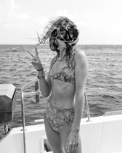 Woman in bikini and diving mask on boat with peace sign in black and white, ocean backdrop. Nautical theme, water sports.