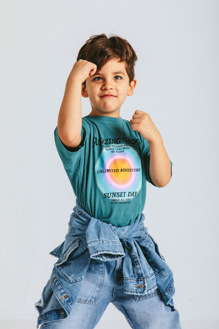 Young boy strikes a playful pose in a turquoise graphic tee, jeans, and denim jacket against a plain white background in a studio setting.