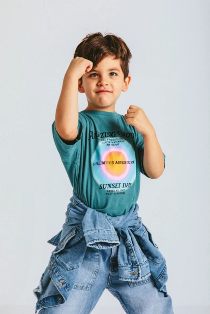 Young boy strikes a playful pose in a turquoise graphic tee, jeans, and denim jacket against a plain white background in a studio setting.