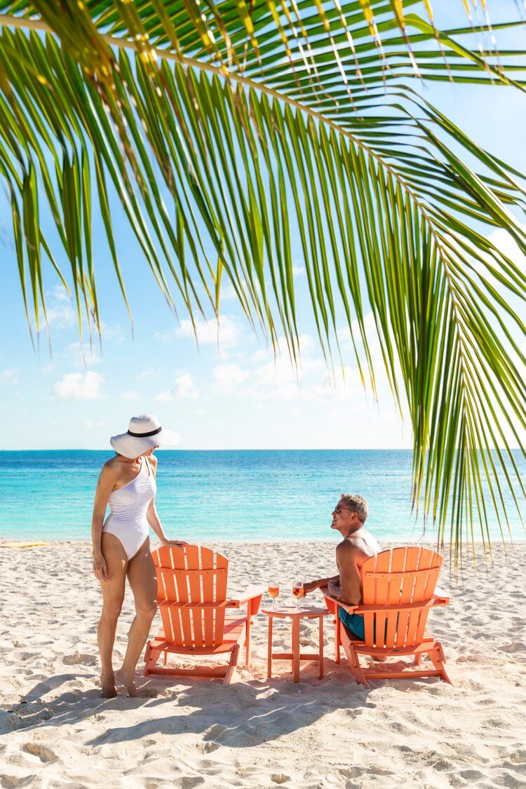 Couple relaxing on tropical beach with turquoise sea. Woman in white swimsuit and hat, man in swim trunks, chairs and table visible.