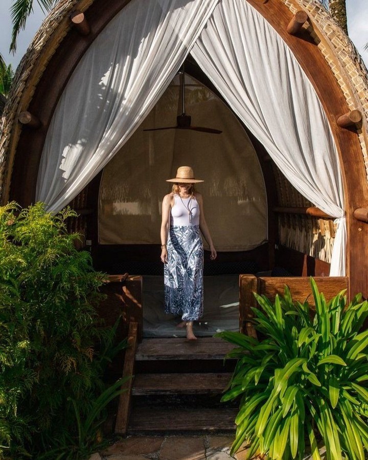 Woman in sunhat and patterned skirt descends from elevated hut with white curtains and tropical foliage. Outdoor, daytime.