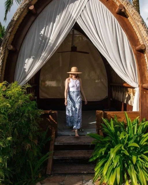 Woman in sunhat and patterned skirt descends from elevated hut with white curtains and tropical foliage. Outdoor, daytime.