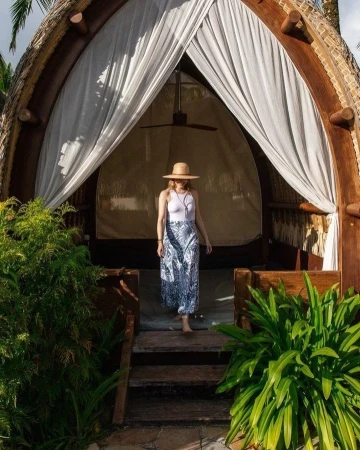 Woman in sunhat and patterned skirt descends from elevated hut with white curtains and tropical foliage. Outdoor, daytime.