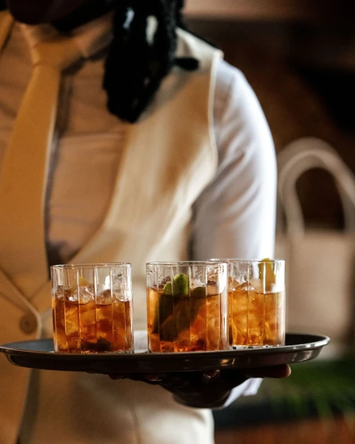 Waiter in formal attire carrying a silver tray with three cocktails garnished with limes, showcasing beverage service.