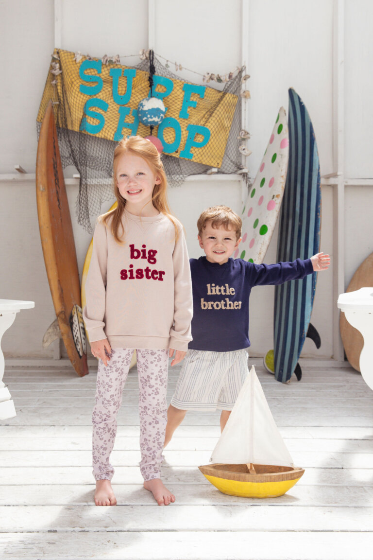 A cheerful girl and boy in matching 'big sister' and 'little brother' outfits pose indoors with surfboards and a toy sailboat.