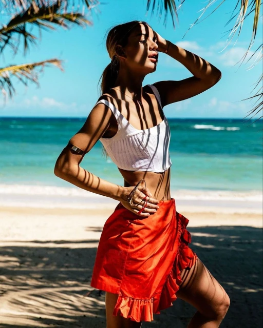 Woman in a white top and orange sarong stands on a sandy beach with turquoise water in the background on a sunny day.