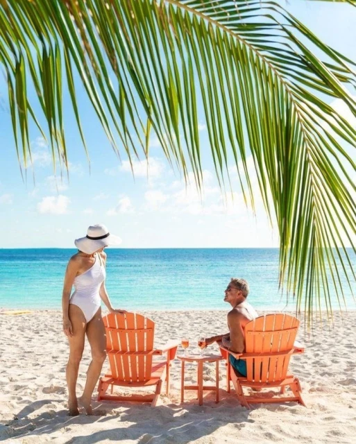 Woman in a white swimsuit and sunhat on a beach next to an orange chair with a man seated and ocean and palm trees in background.