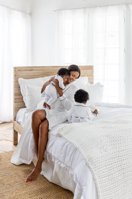 Smiling African-American mother in white robe sits on a bed holding her infant and watching toddler in bright indoor setting.