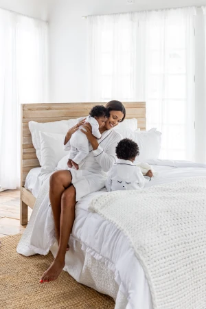 Smiling African-American mother in white robe sits on a bed holding her infant and watching toddler in bright indoor setting.