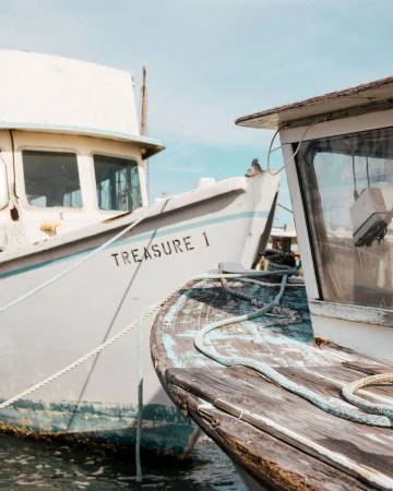 Two docked vintage fishing boats, one labeled 'Treasure 1,' are captured in a close-up shot with cloudy skies in the background.