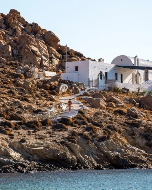 A person in swimwear ascends stone steps toward a whitewashed building on a rocky hillside overlooking turquoise water on a sunny day.