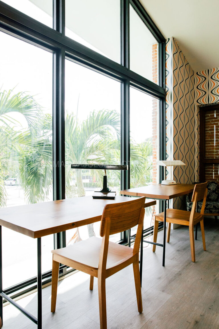 Indoor view with wooden desks and chairs, bright sunlight streaming through large windows, and a vintage-patterned accent wall.