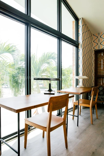 Indoor view with wooden desks and chairs, bright sunlight streaming through large windows, and a vintage-patterned accent wall.