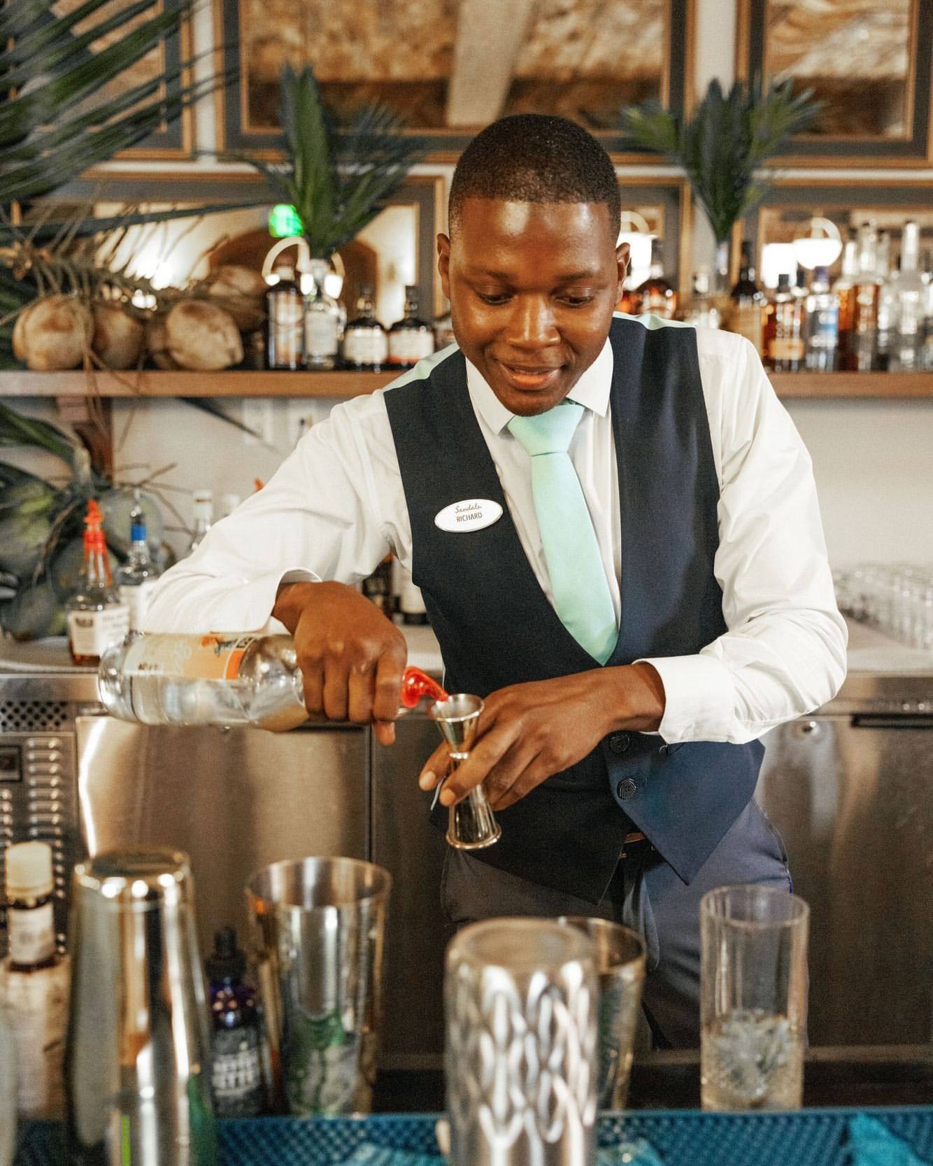 A dark-skinned bartender in a white shirt and vest is pouring clear liquid from a bottle into a metal jigger behind the bar.
