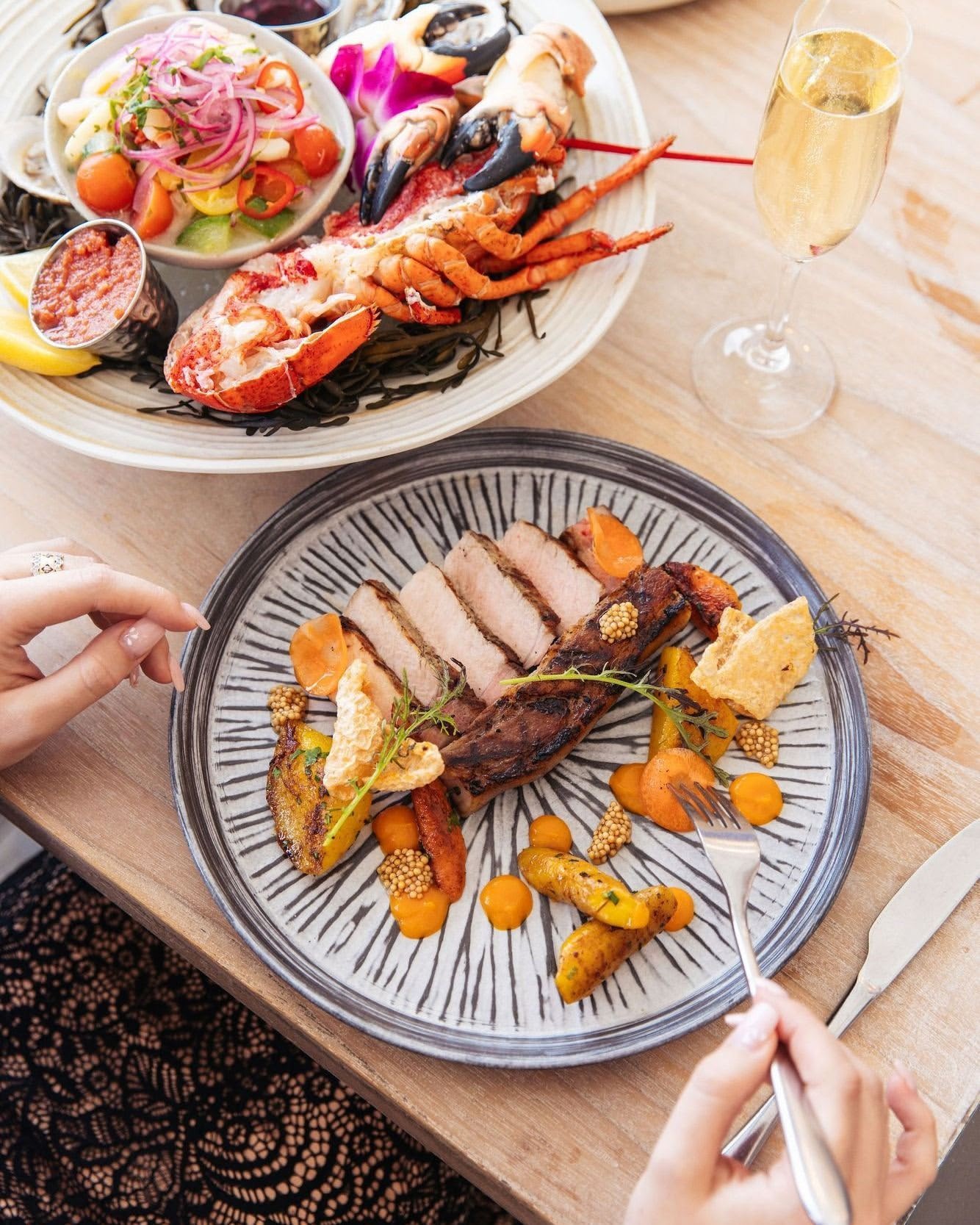 Overhead shot of a woman dining on pork, potatoes, and carrots on a patterned plate, with a champagne glass and seafood platter in view.