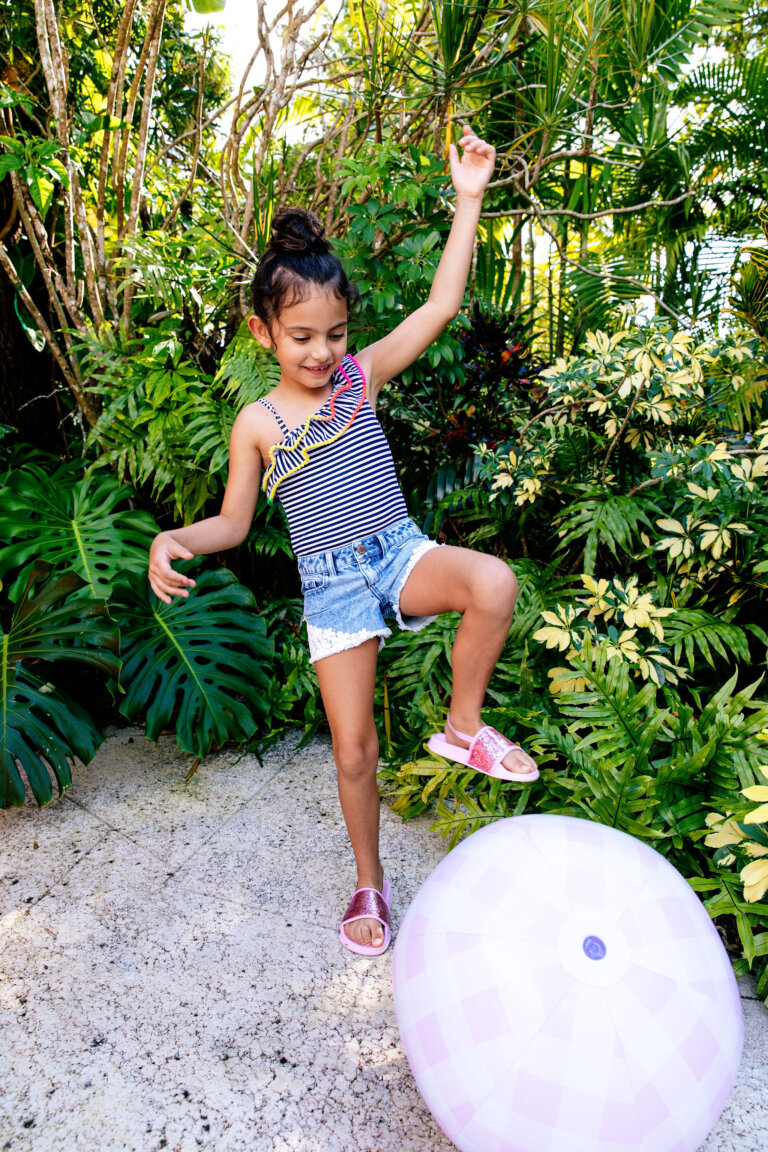 A young girl in a striped swimsuit and denim shorts playfully balances on a large pink ball in a lush, green outdoor setting.