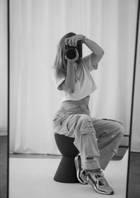 A woman takes a mirror selfie with a digital camera while sitting on a stool in an indoor setting with a white curtain background.