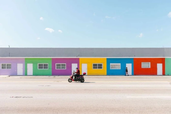 Person riding a red scooter on a city street, with a background of colorful painted buildings under a blue sky, with a second person walking.
