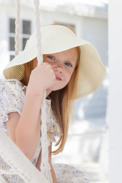 Young girl with red hair and freckles, wearing a wide-brimmed hat, sits on a rope swing in front of a white wooden house.