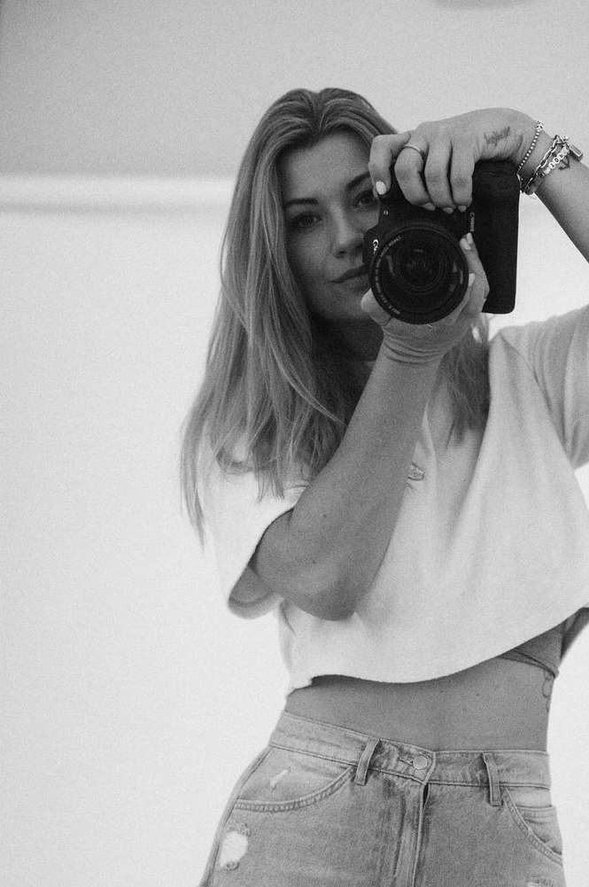 Blonde woman in white top and jeans takes a photo with a DSLR camera indoors, captured in black and white.