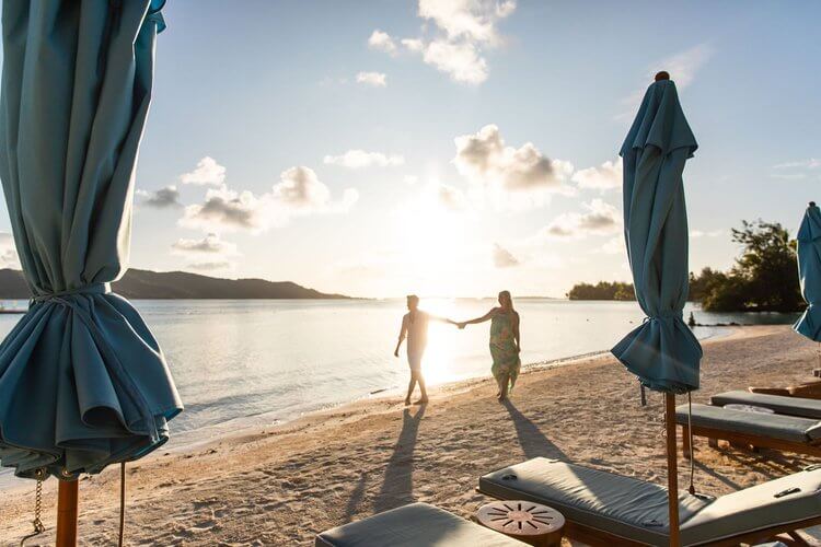 Romantic beach getaway: A couple walks hand-in-hand on a sandy beach at sunset, near turquoise beach umbrellas and lounge chairs.