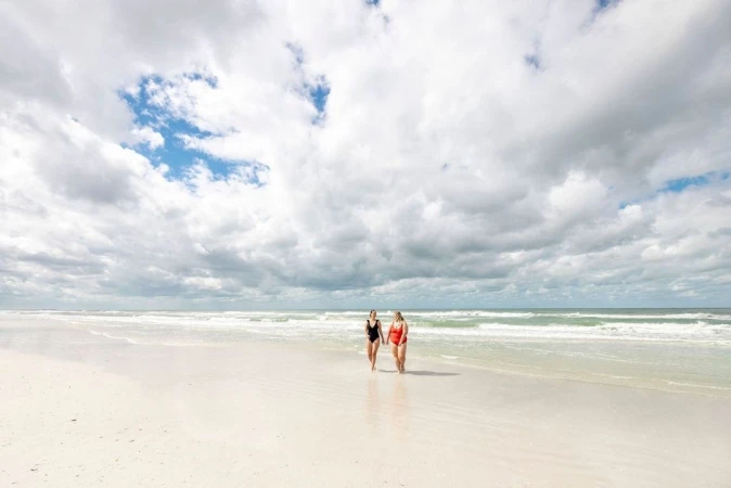 Two women in swimsuits walking together along a wide sandy beach under a cloudy blue sky