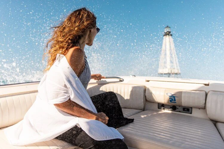 Woman in white shirt and sunglasses rides a powerboat across the sea with a lighthouse and blue sky in the background on a sunny day.