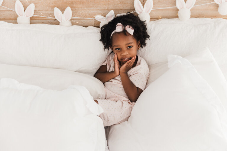Young girl in pajamas sits in a white bed with bunny-ear decorations in the background, looking directly at the camera.
